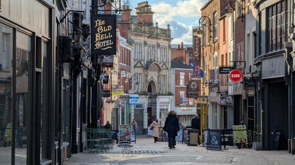 Sadler Gate, a quiet shopping street in Derby. The pedestrianised street winds away down the hill, with a few shoppers walking on the paving stones. On either side of the street there are shops and businesses, including "The Old Bell Hotel", "Bunk" and "Abacus." There are also a couple of "To Let" signs further down the street.