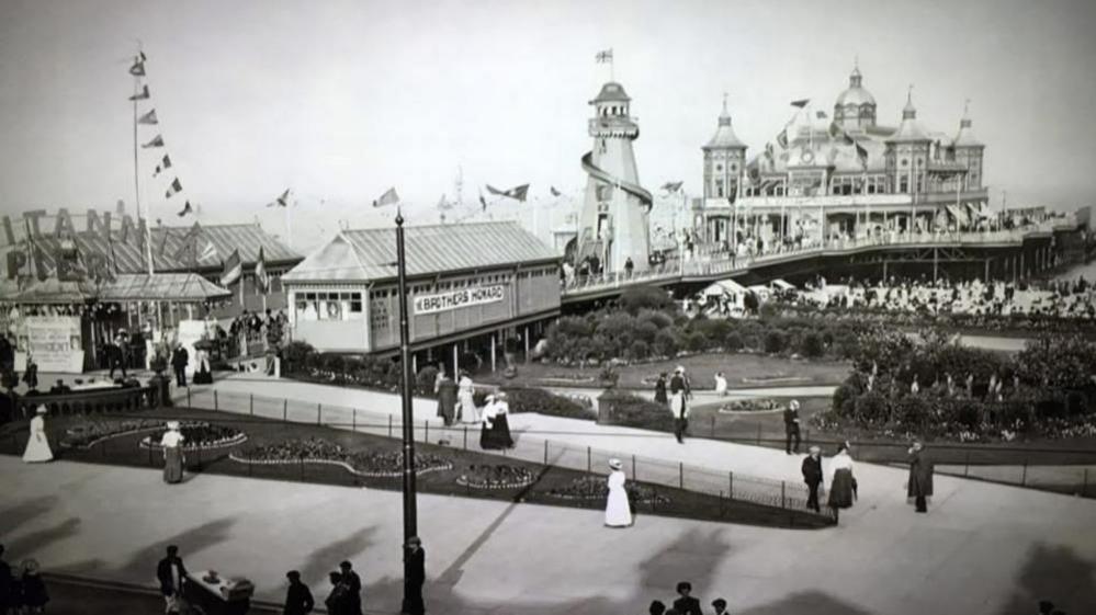 A black and white image of the Britannia Pier as was rebuilt after the fire in 1868. It shows a turreted theatre or pavilion on the seaward end of the building, a helter-skelter and smaller kiosks and booths closest to the landward side.