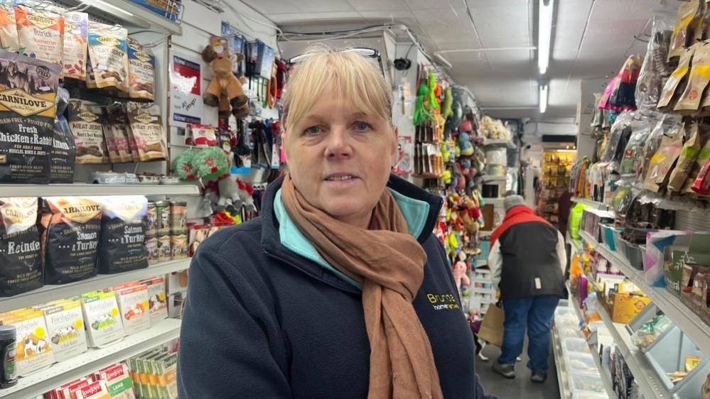 A woman looks into the camera. She has a blonde fringe, and glasses on the top of her head. She is wearing a brown scarf and a dark blue zip-up fleece. She is standing in a shop, and in the background there are products stacked along the wall. 