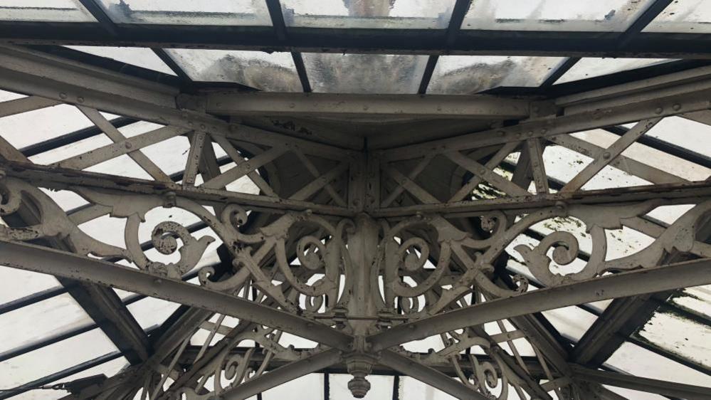 A close up picture of the ornate iron work within the top of the lantern of the Great Yarmouth Winter Gardens, showing the grey sky through dirty glass