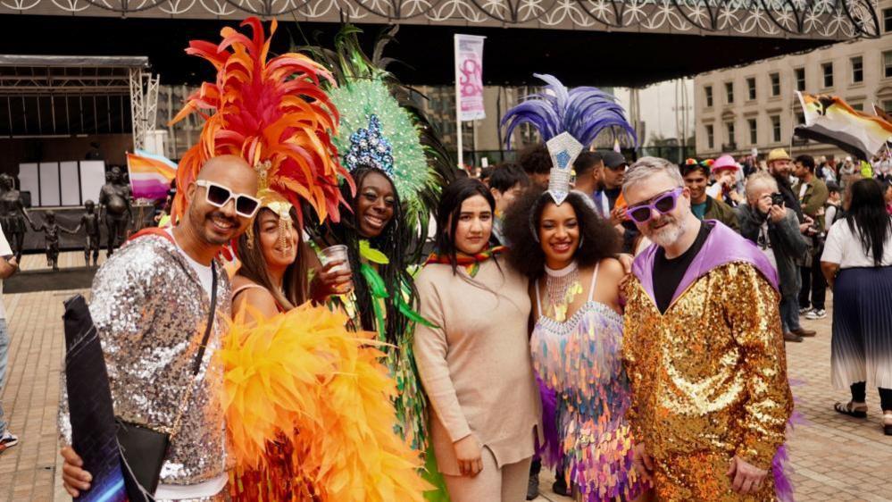 A group of six people standing in front of a large library in a city. They have their arms around each other and are dressed in brightly coloured outfits to celebrate Birmingham Pride. Three of the woman have large feather head dresses and two men are wearing sequin jackets. They are all smiling. 