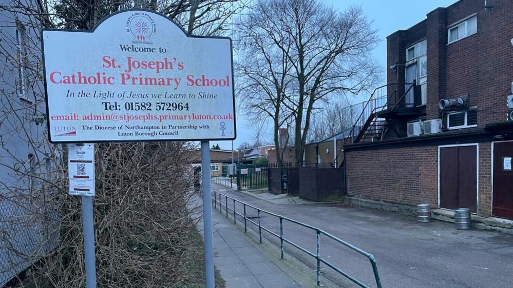 The outside of St Joseph's Catholic Primary School, showing a sign, a walkway, a brick building to the right and a school in the distance. There are metal railings in the middle. 