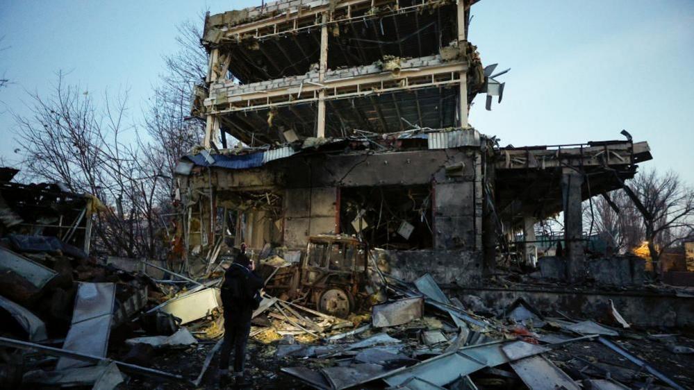 One person stands at the base of a badly damaged building, in front of which is a burned out tractor and large piles of debris