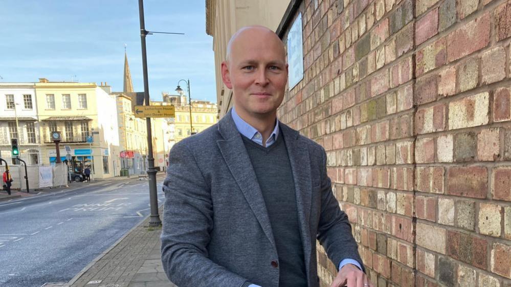 Liberal Democrat MP for Cheltenham, Max Wilkinson in Cheltenham February 2024. He is smiling at the camera and is wearing a grey jumper, grey jacket and a light blue shirt