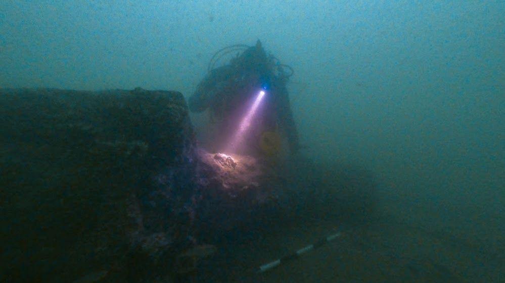 A scuba diver shining a torch on an old warship cannon on the seabed. Around them the sea is different shades of green