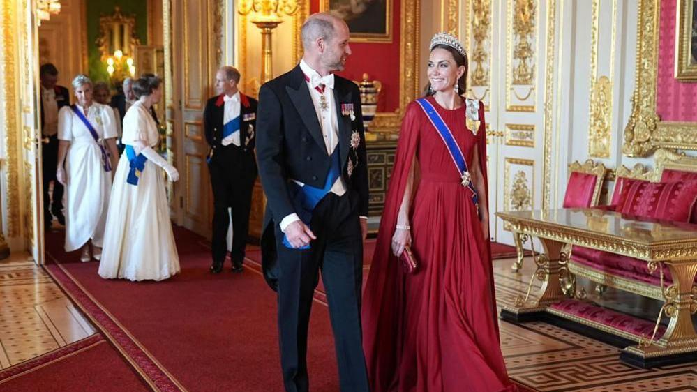 The Prince and Princess of Wales smile at each other as they walk into the state banquet. They are followed by other senior royals who are chatting to each other as they walk through a door.