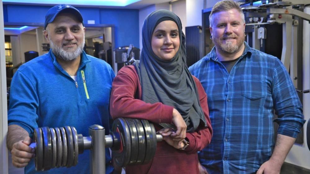 Two men and a woman stand in a gym. The man on the left and the woman in the centre lean on a rack with weights on it. 