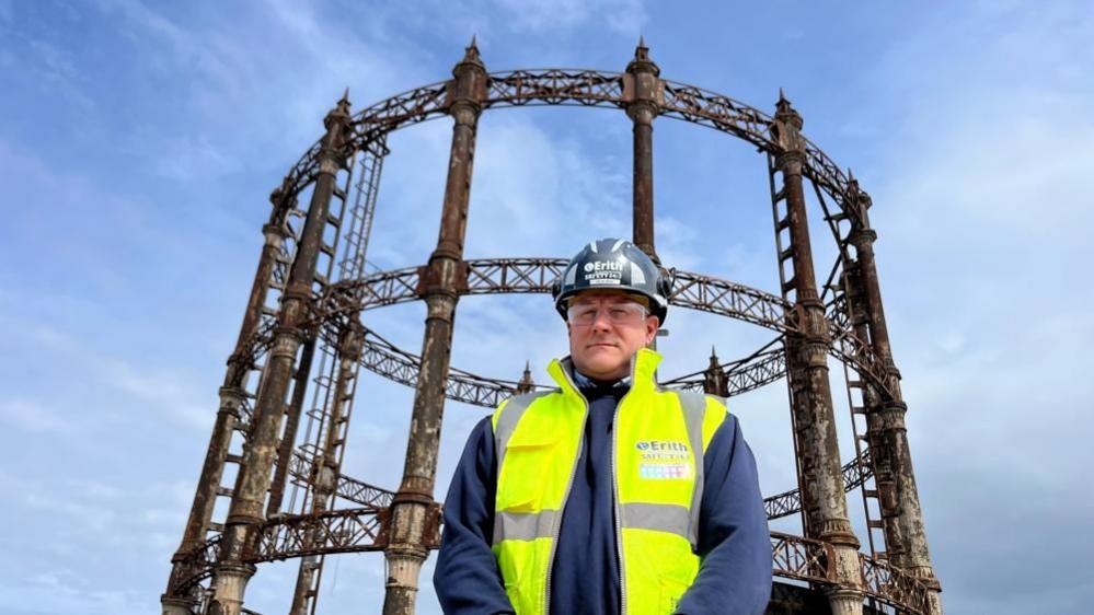 Gavin Craig is standing in front of the Great Yarmouth gasometer, wearing a white hard hat, safety glasses, and a high visibility yellow tabbard over his blue jacket. Behind him is the gas holder, which is made from cast iron columns and lattice work, with finials on top of the columns. The sky behind is a grey/blue.
