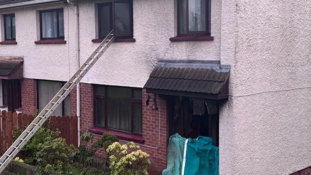 A ladder is leading to the upstairs window of a burnt out house. A green protective sheet is covering the front door.