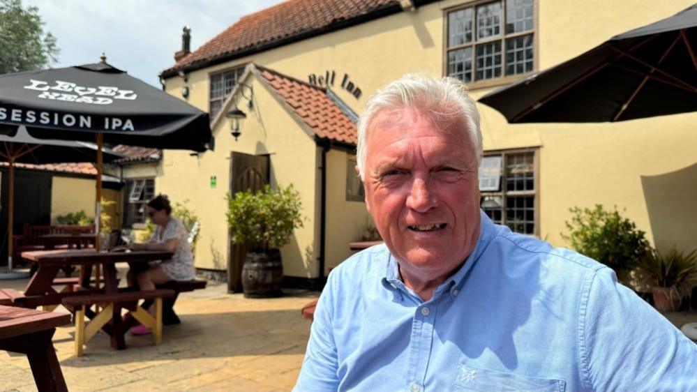 David Moss is wearing a blue shirt. He has white receding hair, and is sitting at a table outside The Bell Inn in Carlton Colville, of several pubs he operates. The building is rendered, and painted a ochre yellow. There are other tables nearby, with black parasols above them.