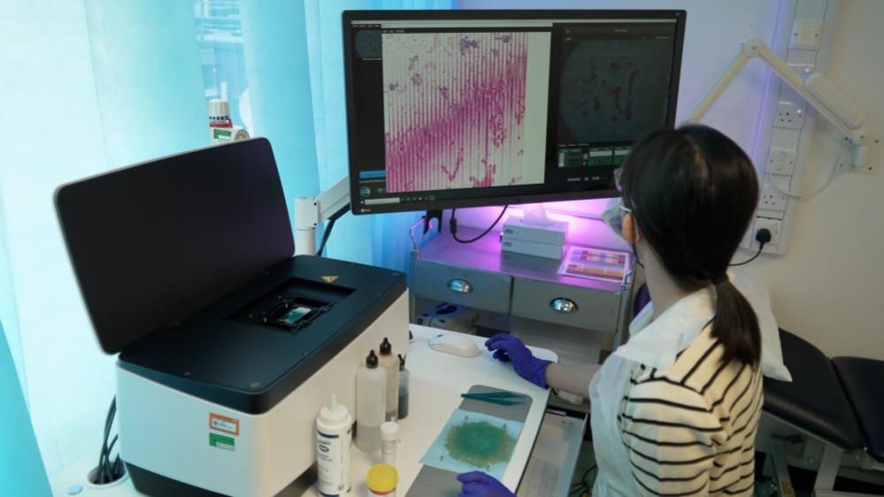 A woman is looking at a computer screen in a laboratory. She is wearing goggles and has dark haired pulled back in a pony tail. In front of her is a scanner and various bottles probably containing chemicals