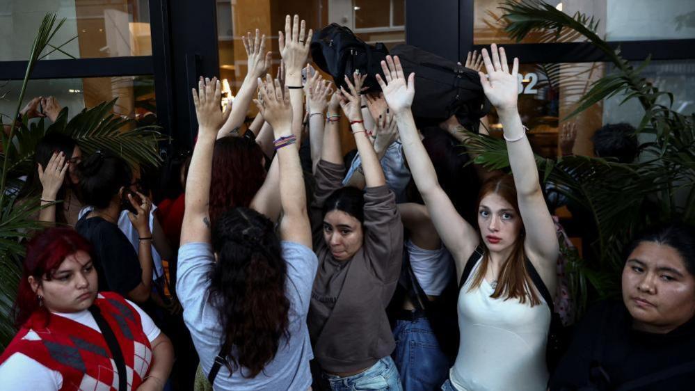 A group of mostly female fans hold their arms high in the air outside the front door of Casa Sur hotel in Buenos Aires, Argentina. They are trying to block the view of cameras as Liam Payne's father, Geoff, visits the hotel where his son died. Geoff Payne cannot be seen in view.