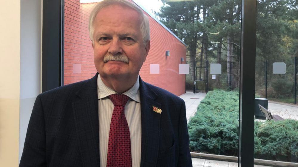 A staff member of the police force in a suit and red tie. He is 70 years old and is standing in front of a full length glass window where you can see the outside. There are trees and bushes. 