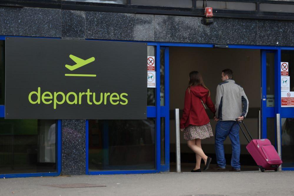 Woman wearing red coat and dress with man in grey jacket pulling red suitcase through blue sliding door with sign reading 'departures' beside it. 