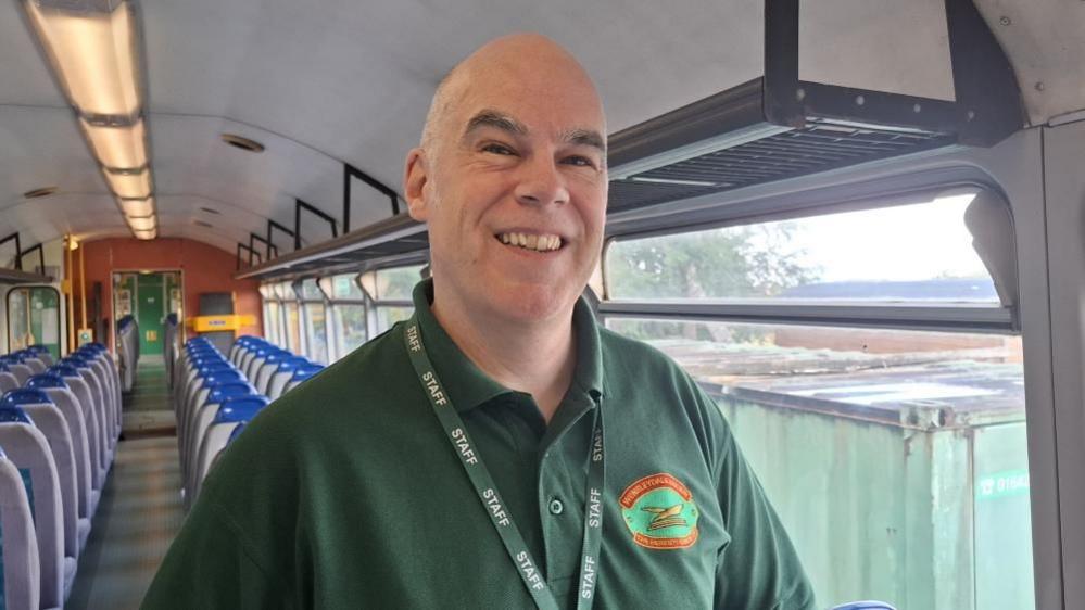 A man smiles at the camera, standing in a railway carriage. He is wearing a dark green polo shirt with the Wensleydale Railway logo on it, and a lanyard which reads "staff".