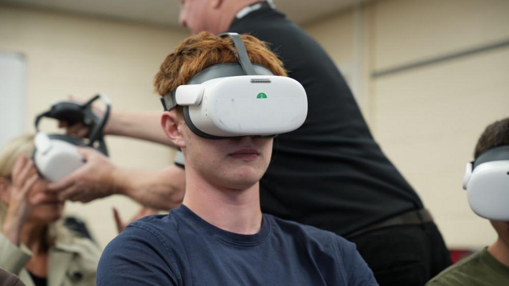 A young man with red hair and wearing a blue T-shirt wears a pair of virtual reality goggles. People can be seen blurred in the background.