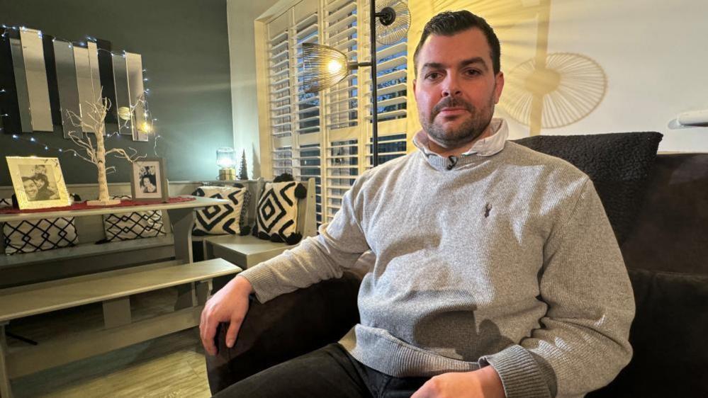 A man in a grey jumper, over a white shirt, sitting on a sofa in his lounge, with family pictures in the background on a table. 