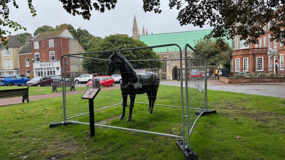 A fibreglass black horse, painted with images of Black Beauty and author Anna Sewell, sited with an information board, outside Great Yarmouth Minster and the house where Ms Sewell was born, which is out of vision in this image. The image shows the horse leaning to its left. Behind it are other buildings.