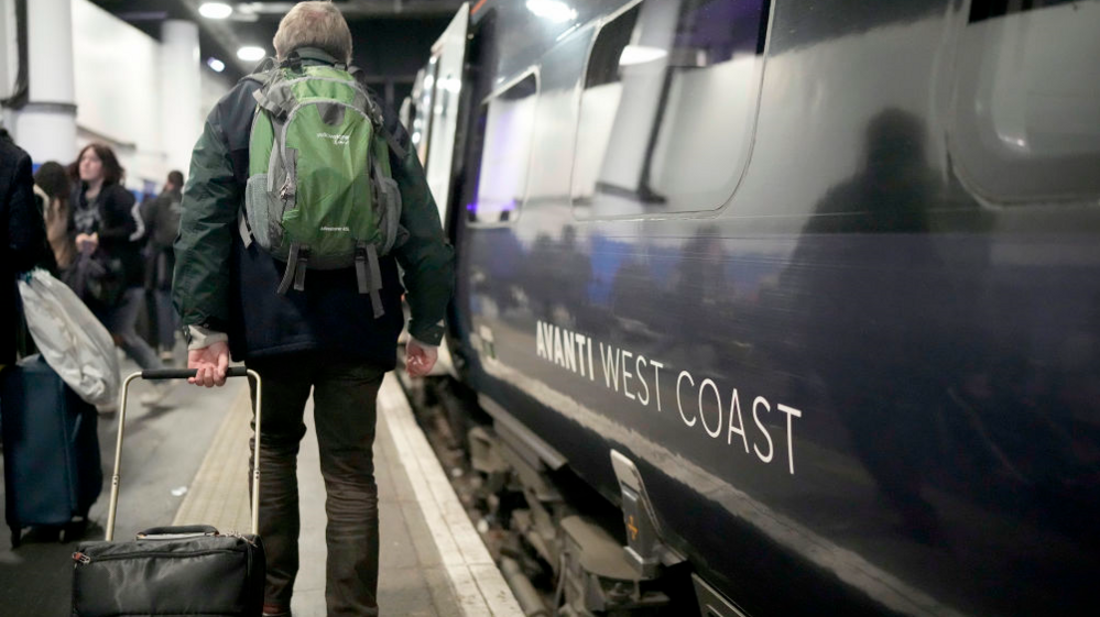 Man in dark clothes with a green rucksack and wheeled suitcase walks alongside an Avanti West Coast train  