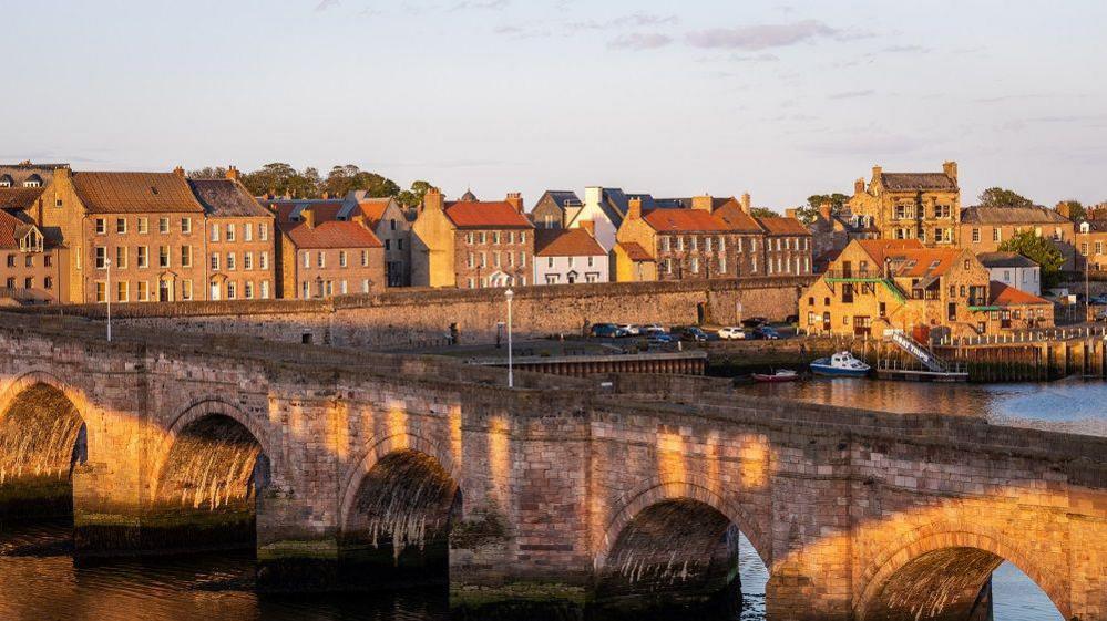 A photo of Berwick from one side of a bridge. The buildings are all made from stone and the river sits in front of them. The stone bridge is in the forefront of the photo with a river running underneath a series of arches.
