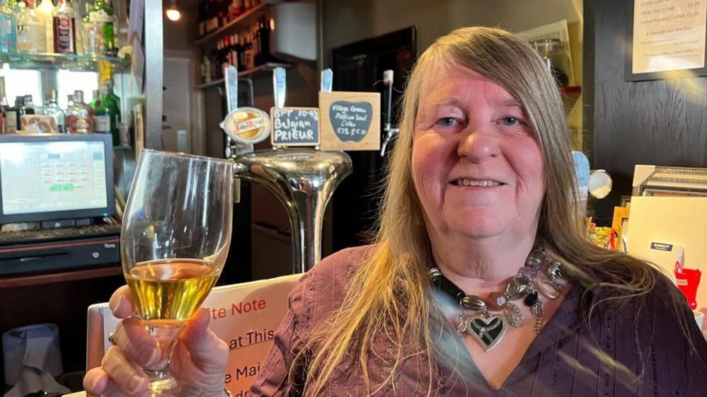 Sarah Newson has long blonde hair, is wearing a necklace and has a purple v-neck top. She is holding a glass of cider, near the bar of the Blackfriars Tavern.