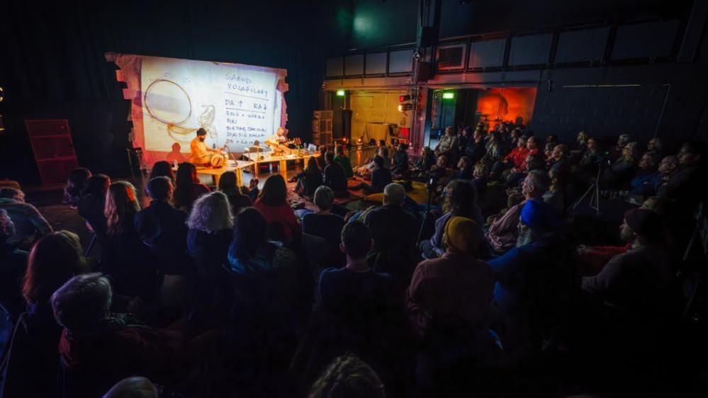 A performance at the New Sounds South Asian Music festival in Bristol in 2023. Dozens of people are sat on the floor in the dark watching a performer on a lit stage. 