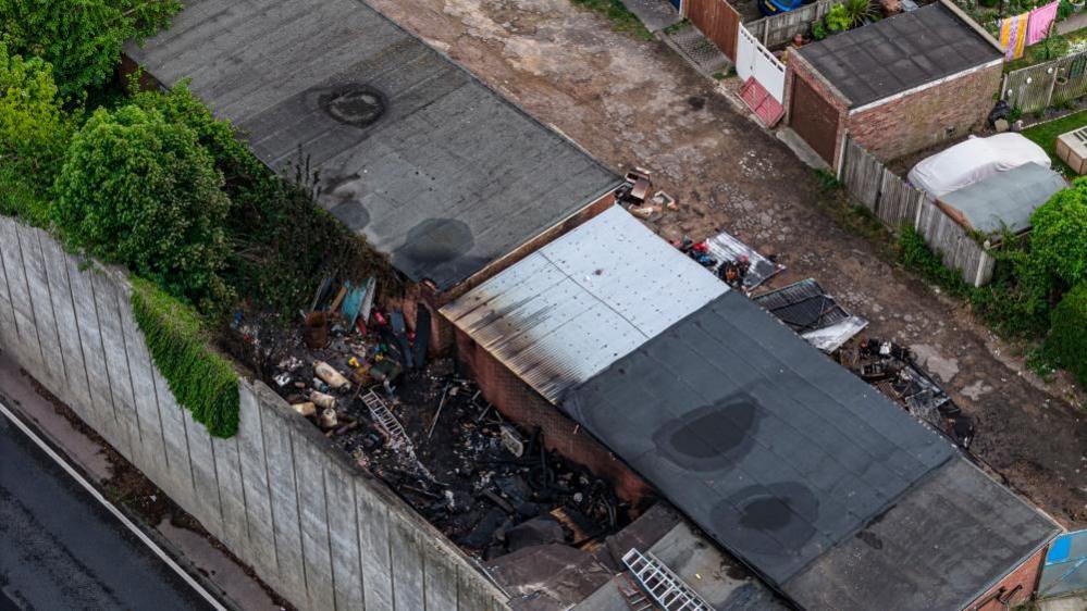 A drone image shows a row of garages, with burned and charred debris either side of the building. One of the garages has a corrugated iron roof, where the others have felt roofs. None has been destroyed by the fire. A concrete retaining wall, with the A47 below, can be seen in the lower left of the image. Other garden buildings and garages can be seen in the upper right of the image.