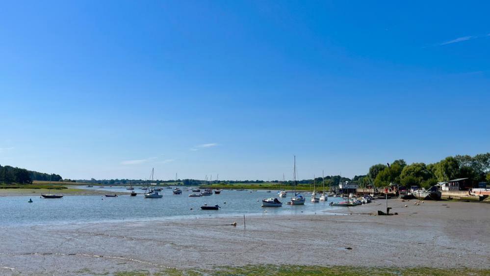 A picture of the River Deben from the quayside in Woodbridge.  The sky is big and blue the tide is out so there some mud flats and then the river. On the rivers there are some boats bobbing in the distance. 