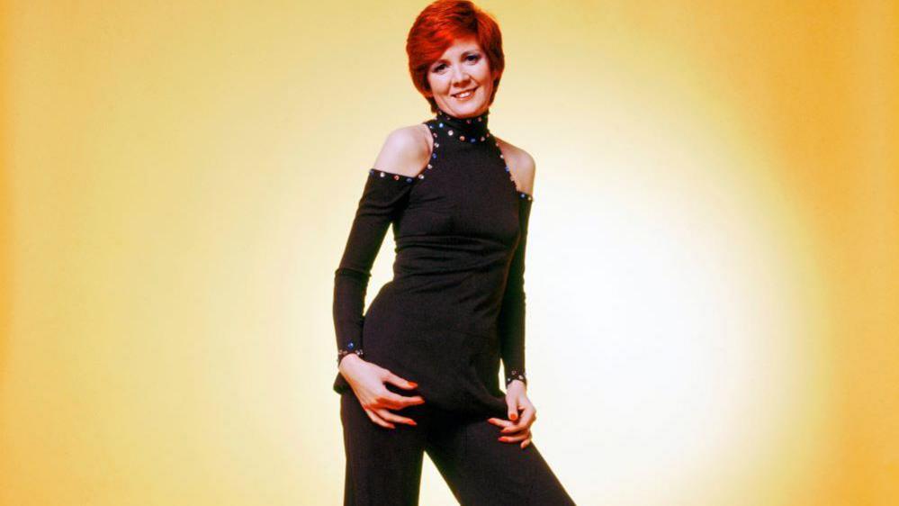 A young Cilla Black poses in front of a yellow background. She wears a black jumpsuit with cut out shoulders and a high neck. She has short red hair.
