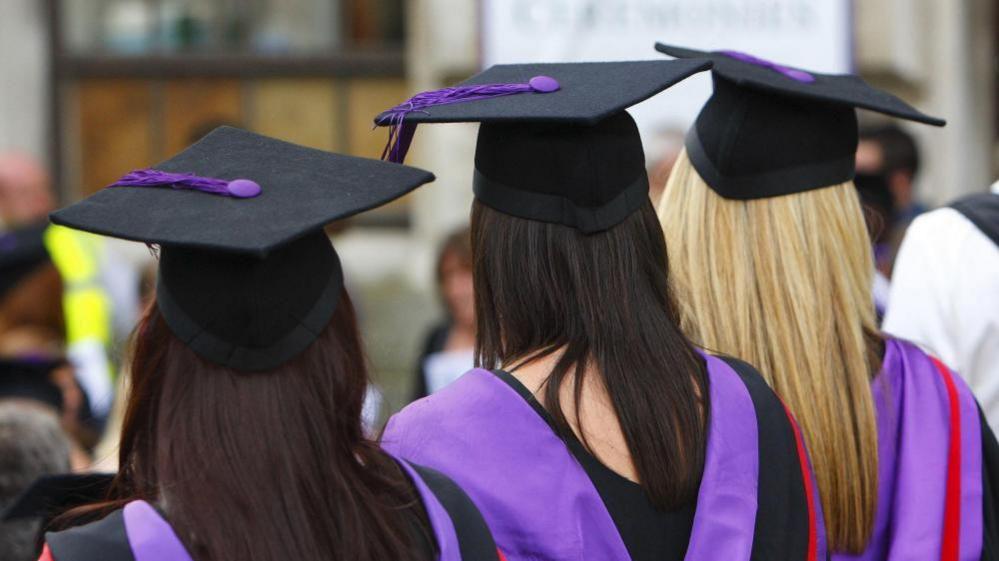 Students wearing a black graduation cap. They have long hair and a black gown with purple detailing.