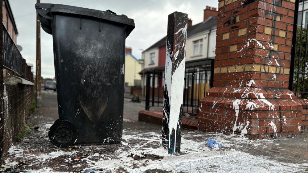 Black wheelie bin on pavement, covered with white paint, red brick wall graffitied 