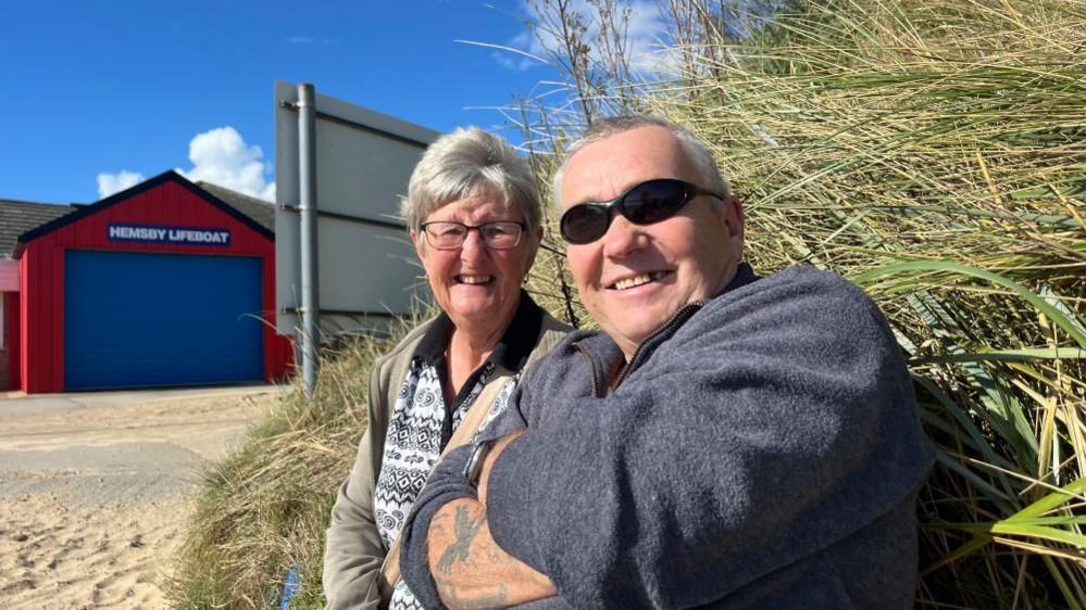 John Peckham in a blue top with sunglasses and his wife Vanessa in a patterned top, sitting near the marram grass, with Hemsby Lifeboat Station in the background