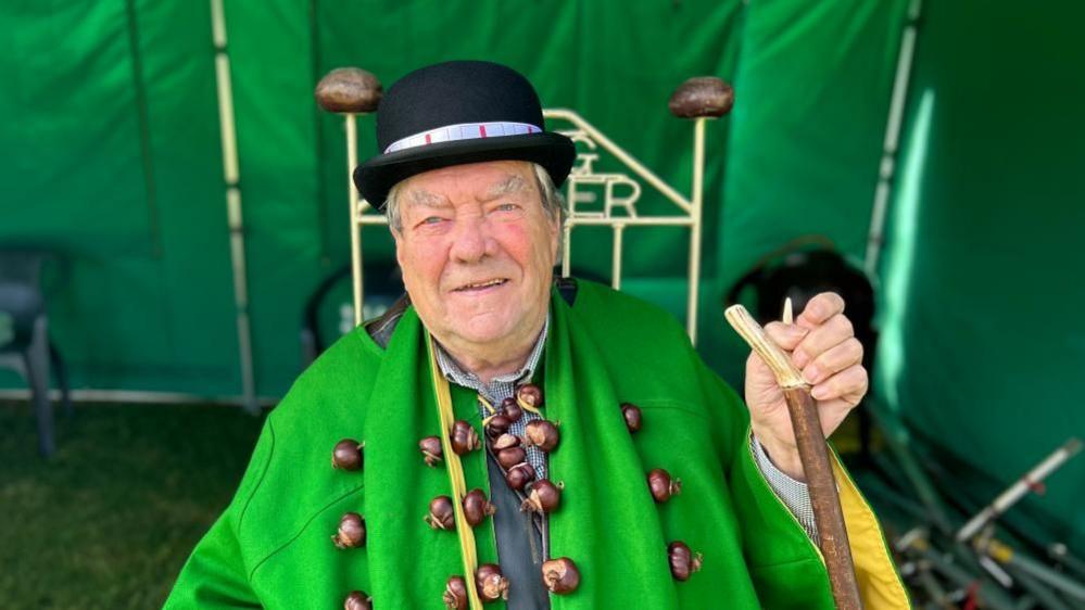 David Jakins with short white hair wearing a black bowler hat and a green cloak decorated with conkers. He is holding a wooden stick and sitting in a green marquee.