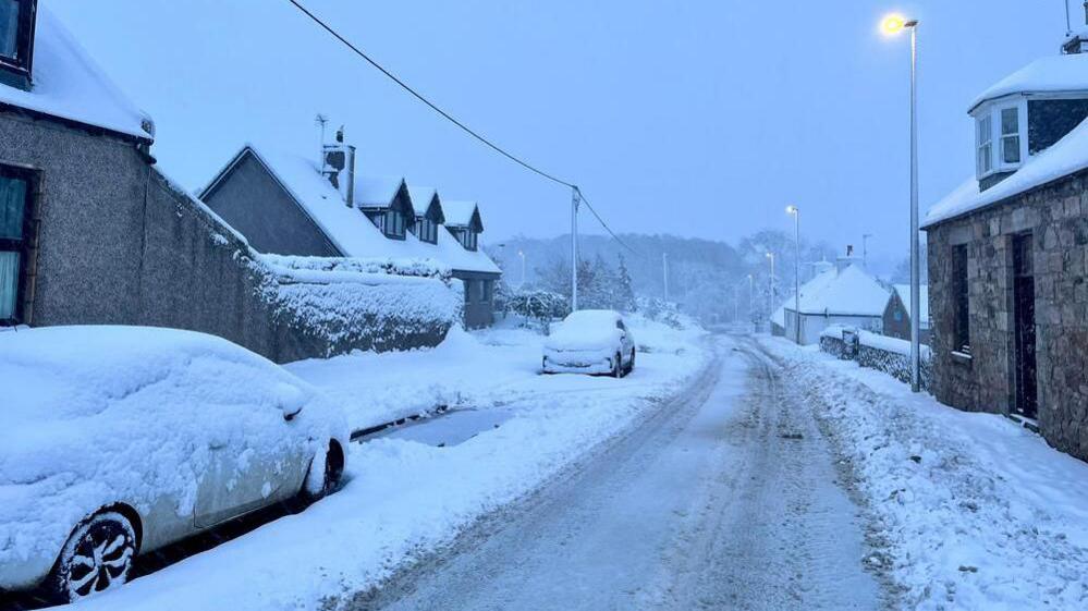 Deep snow covers cars and houses on a cloudy morning. A path has been worn through the road by cars.