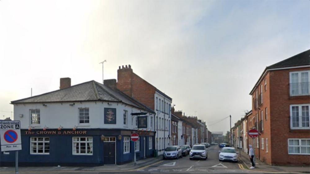 A two-storey pub building with traditional swinging sign saying "Crown and Anchor". The name of the pub is also above the windows on the ground floor. The ground floor is painted blue and the upper floor is white. There are cars parked on both sides of the adjacent street of terraced houses.