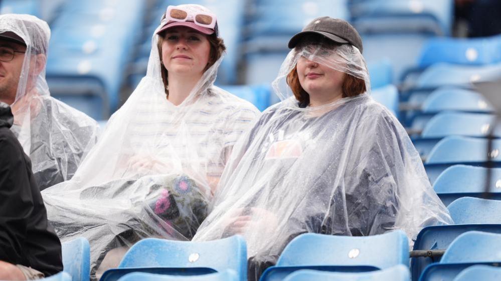 Two young women wearing caps and plastic ponchos are seen smiling as they look on from the stands at a cricket game in Leeds on Wednesday
