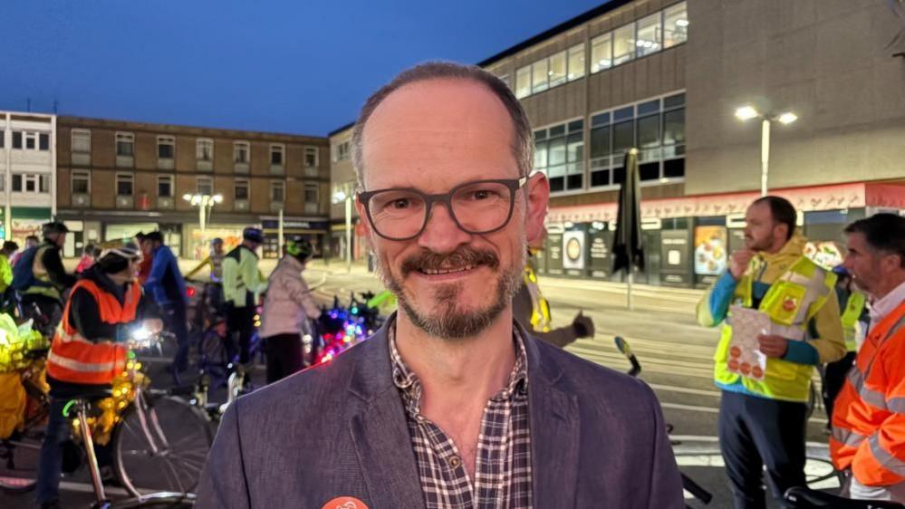 Alistair Gordon with short brown hair and beard and glasses. He is wearing a purple jacket over a red, grey and white checked shirt. He is standing in a square with cyclists in the background and three-storey shop and apartment buildings in the background.