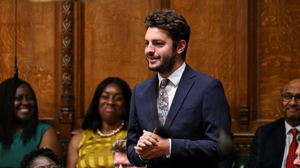 Jack Abbott speaking in Parliament. He has black hair and a beard and is wearing a dark blue suite with a white shirt and a patterned tie. He has his hands clasped in front of him. Three other MPs can be seen sitting on the bench behind him. There is wooden panelling behind them.