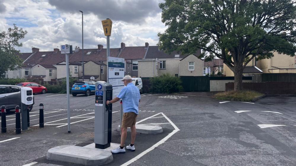 Wearing brown shorts, a blue short sleeved shirt and a flat cap, a man can be seen paying for parking in a Kingswood car park.