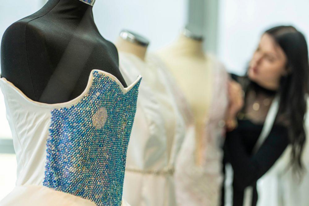 A close up picture of a blue sparkly bodice of a dress on a mannequin. Two mannequins are behind and a woman is working on one of the dresses.