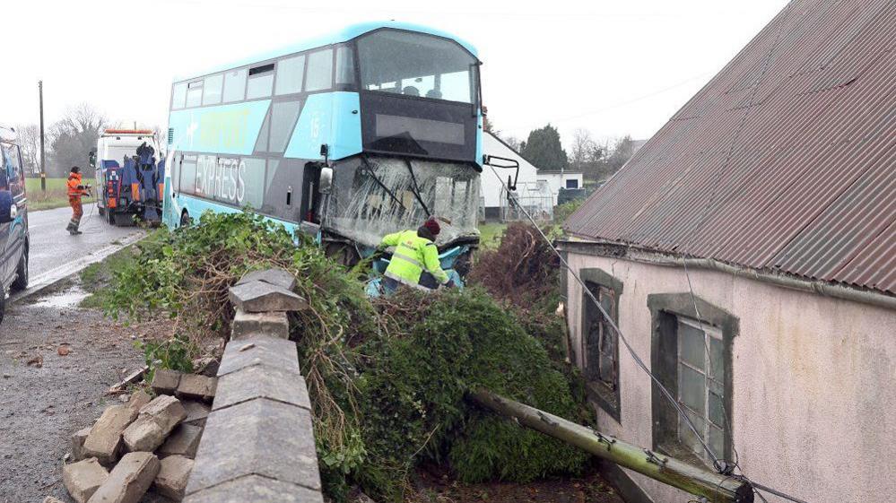 Large blue bus with smashed windscreen, crashed into a wall, men in Hi Viz working on road, smashed wall and large tree lying on ground