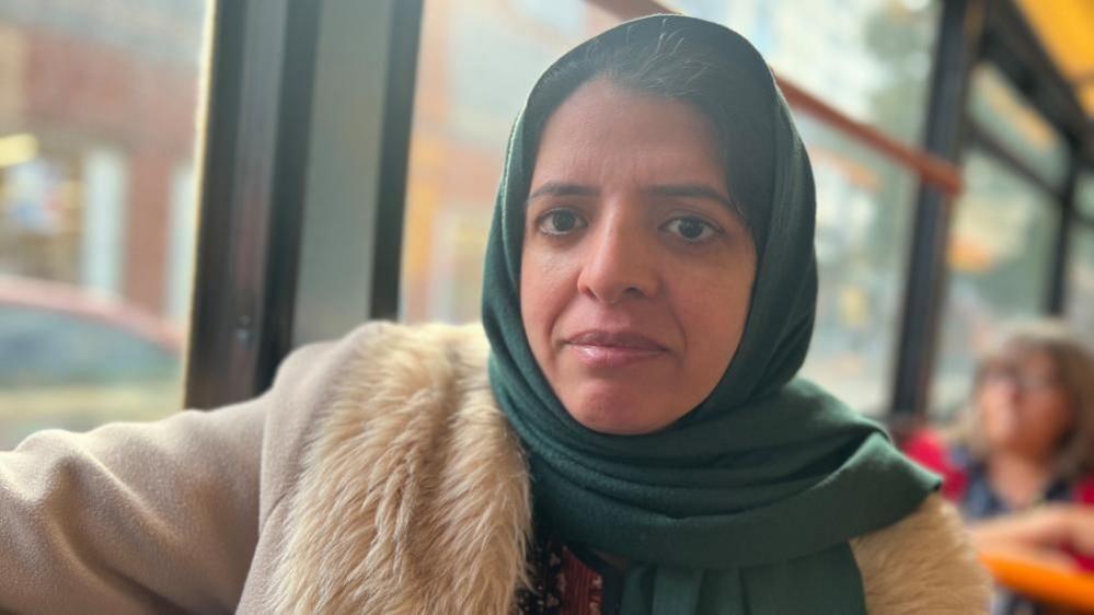 Maria, a woman wearing a green headscarf and wearing a cream coat, sitting on a bus and looking at the camera 