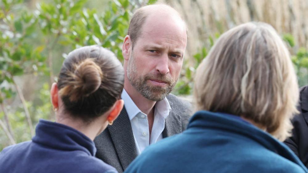 The Prince of Wales wearing a grey suit looks towards the camera while two staff face him. 