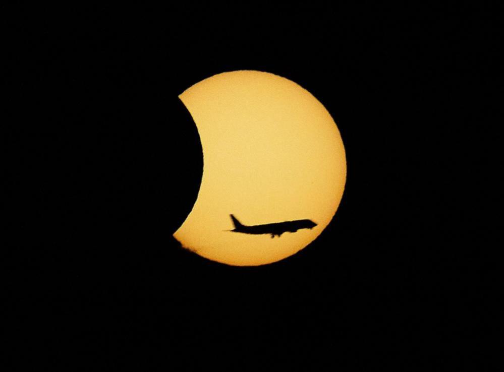A plane flies in front of the Moon passing the Sun, which is a large yellow circle against the black sky 