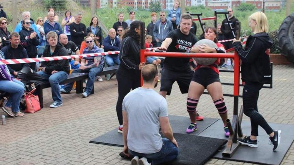 Louise Clark in black shorts and a red t-shirt, lifting a huge heavy grey cement ball as part of the Strong Women Contests in 2016.