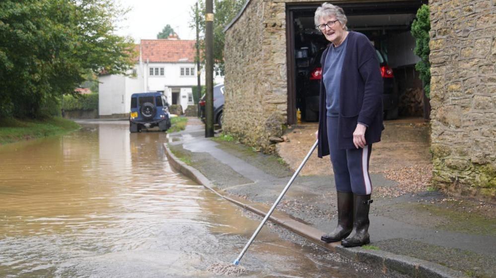 A woman with white hair wearing a blue cardigan is standing on the pavement and pushing a mop into a flooded road.  There is a stone wall behind her and a Land Rover is seen in the distance.