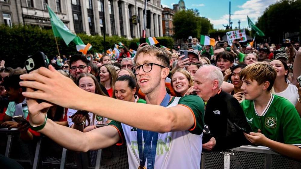 Gold medal winning swimmer Daniel Wiffen, wearing an Ireland top, takes a selfie in front of the crowd in Dublin with Irish flags being waving in  the background