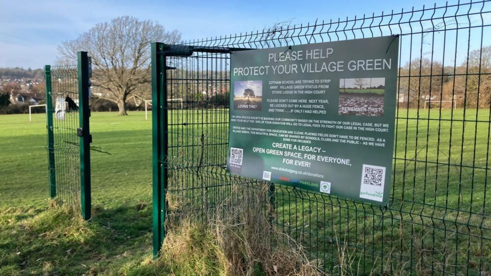 A green field, with a fence and a sign posted on it that reads "please help protect your village green"