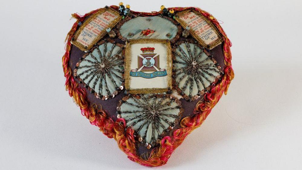 A heart-shaped cushion with lots of decoration, including army regiment badge, phrases and sequins.