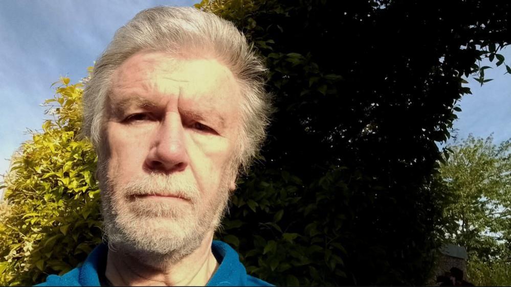 David Morgan with grey hair and beard wearing a blue top in a garden